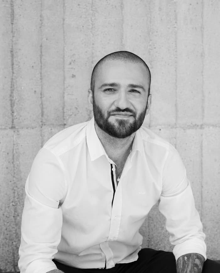Amin Masoudi, assis devant un mur de béton, chemise blanche, sourire discret